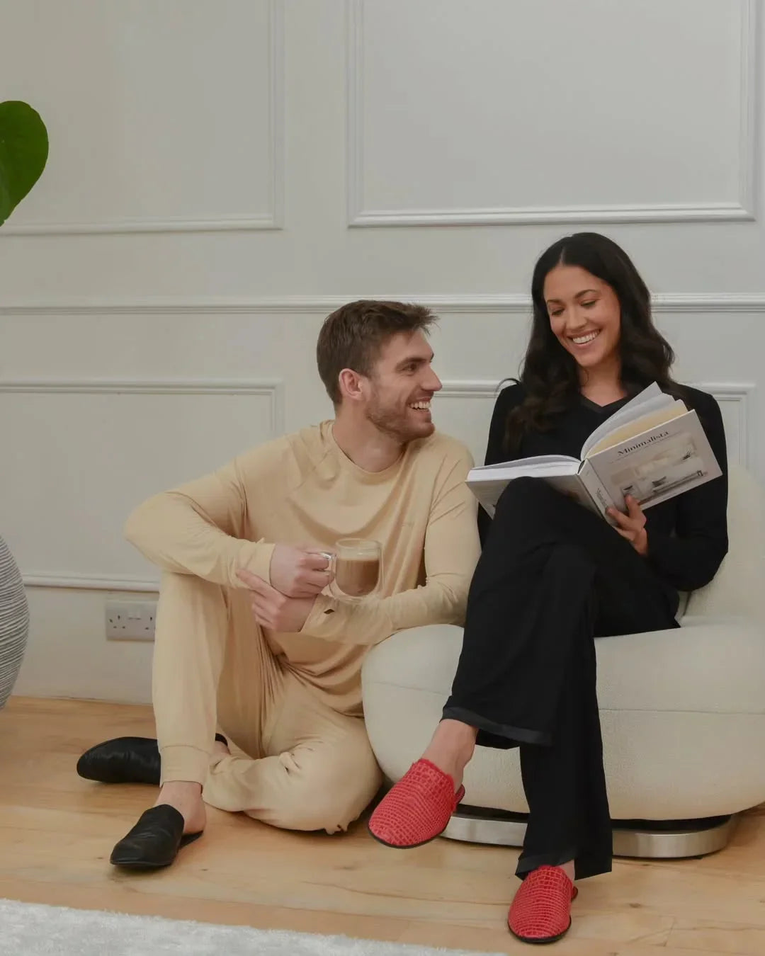 Couple relaxing indoors, man with coffee and woman reading, both in cozy loungewear and slippers